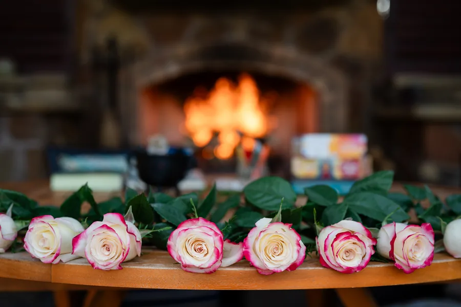 A wood fire in a stone hearth, with cream-and-pink roses laid out on a wooden table in front, a small cauldron and oracle decks in between