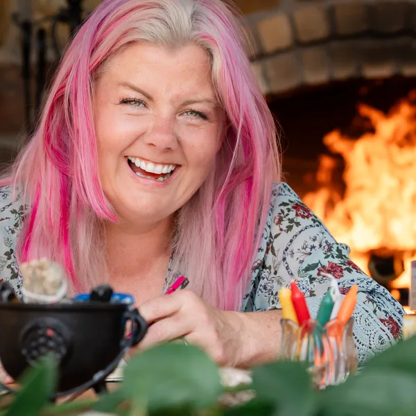 Erin Nes laughing in front of a fireplace, pink hair, holding a cauldron with her oracle decks beside her
